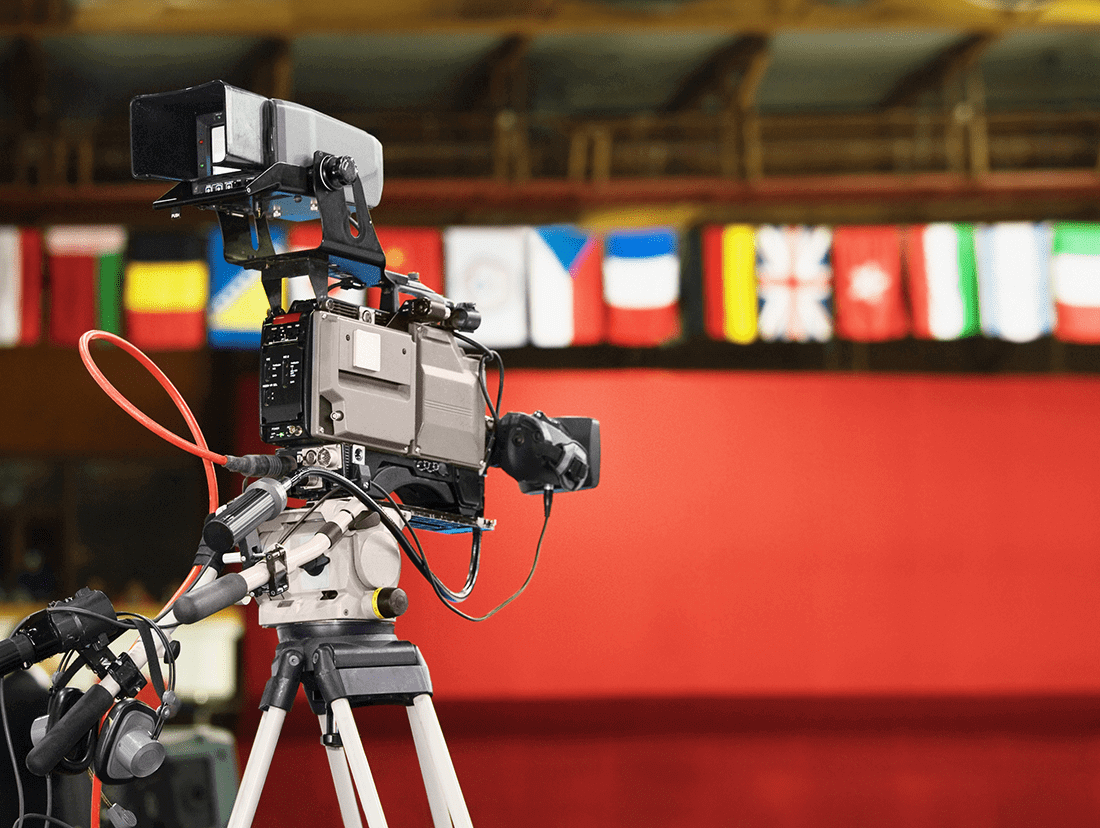 A video camera on a tripod in front of flags.